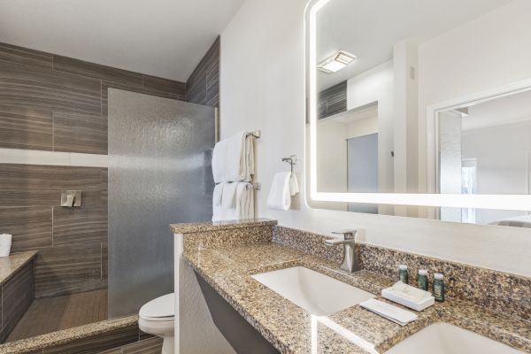 The image shows a modern bathroom with a double sink vanity, granite countertops, a large mirror with lighting, towels, and a walk-in shower.