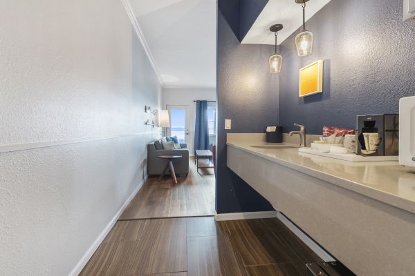 A hallway with a kitchenette on the right, leading to a living area with a sofa, coffee table, and curtains drawn to one side at the window.