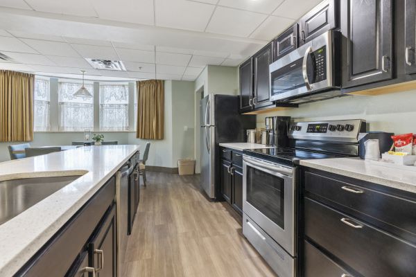 A modern kitchen with stainless steel appliances, dark cabinets, a microwave, a coffee maker, and a dining area with windows and curtains.