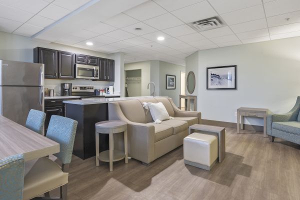 The image shows a modern living area with a sofa, kitchen, and dining space, featuring sleek furniture and decor in light and neutral tones.