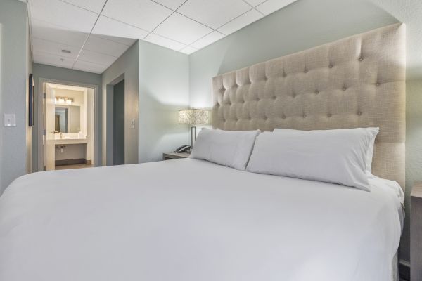 The image shows a neatly made bed with white linens in a modern hotel room, featuring a tufted headboard and a view leading into a bathroom.