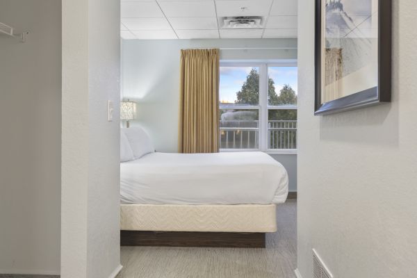 The image shows a modern hotel room with a neatly made bed, a window with yellow curtains, a bedside lamp, and artwork on the wall.