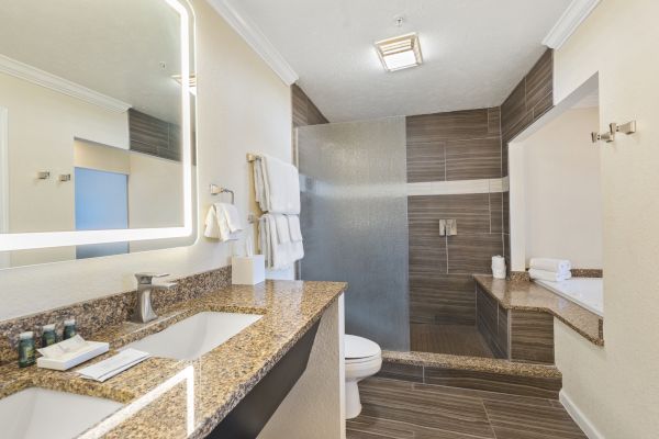 A modern bathroom features a double sink vanity, backlit mirror, stacked towels, a toilet, a walk-in shower with frosted glass, and a large bathtub.