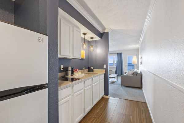 The image shows a narrow hallway leading to a living area. On the left, there's a small kitchen with white cabinets and pendant lights, and a fridge.