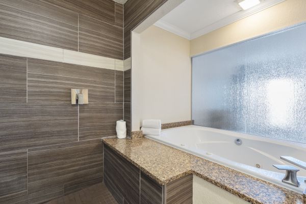 A modern bathroom with brown tiled walls, a granite countertop, a white bathtub, a frosted window, and a soap dispenser.