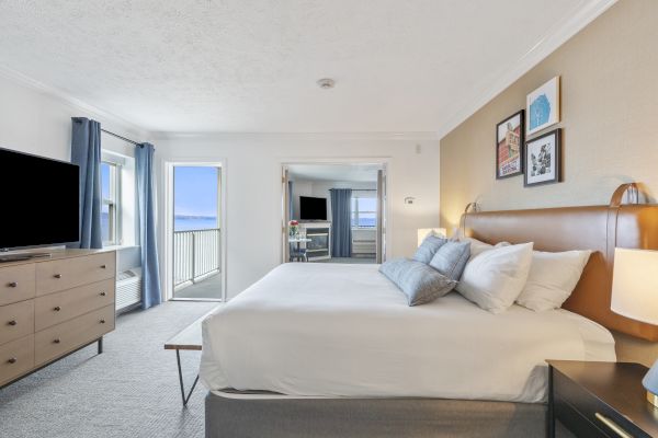 A bright bedroom features a large bed, dresser, TV, balcony with ocean view, and modern decor on the walls, creating a cozy atmosphere.