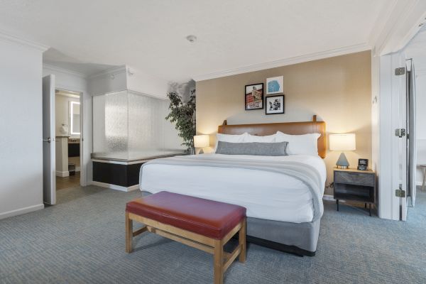 This image shows a hotel room with a large bed, two bedside tables with lamps, a bench at the foot of the bed, and an adjacent bathroom.