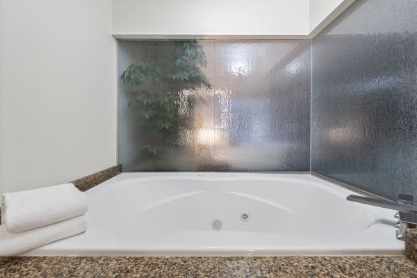 A white bathtub with a towel on the side, granite countertop, and frosted glass window with a view of a plant and lamp in the background.