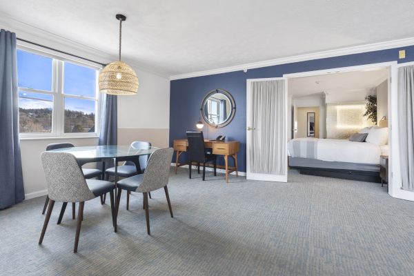 A modern hotel room with a dining table, four chairs, a writing desk, a wall mirror, and a separate bedroom area with a bed and white linens.