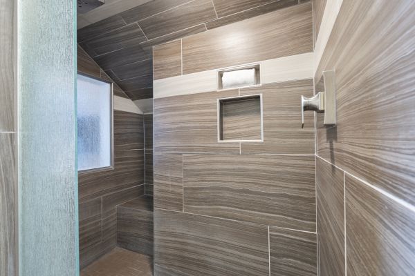 The image shows a modern tiled shower with a bench, a recessed shelf, and a frosted window, featuring gray and beige tones arranged in a contemporary pattern.