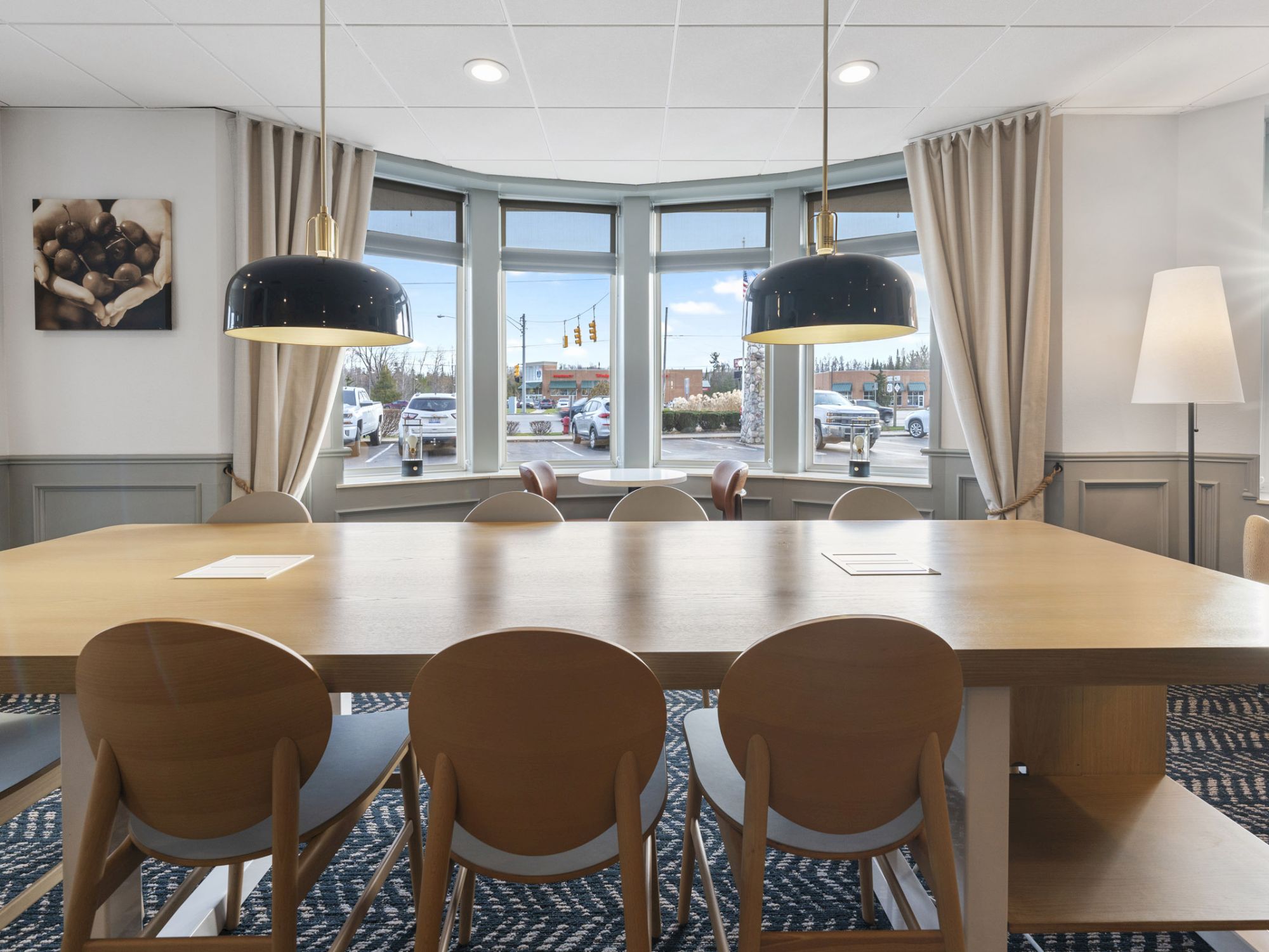 A modern meeting room with a long wooden table, several chairs, pendant lights, large windows, curtains, and a floor lamp. It has a clean, bright look.