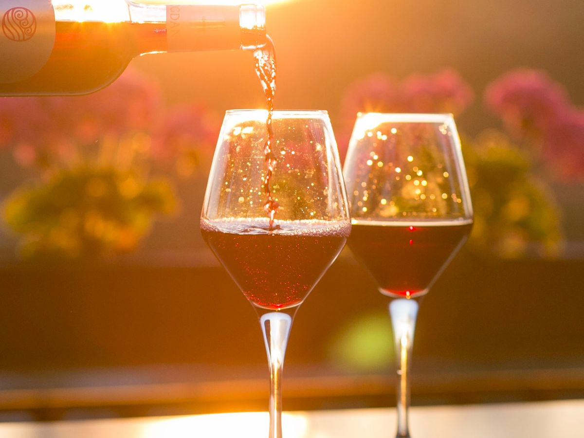 A bottle of wine is being poured into two glasses, with a scenic sunset background and flowers in the distance.