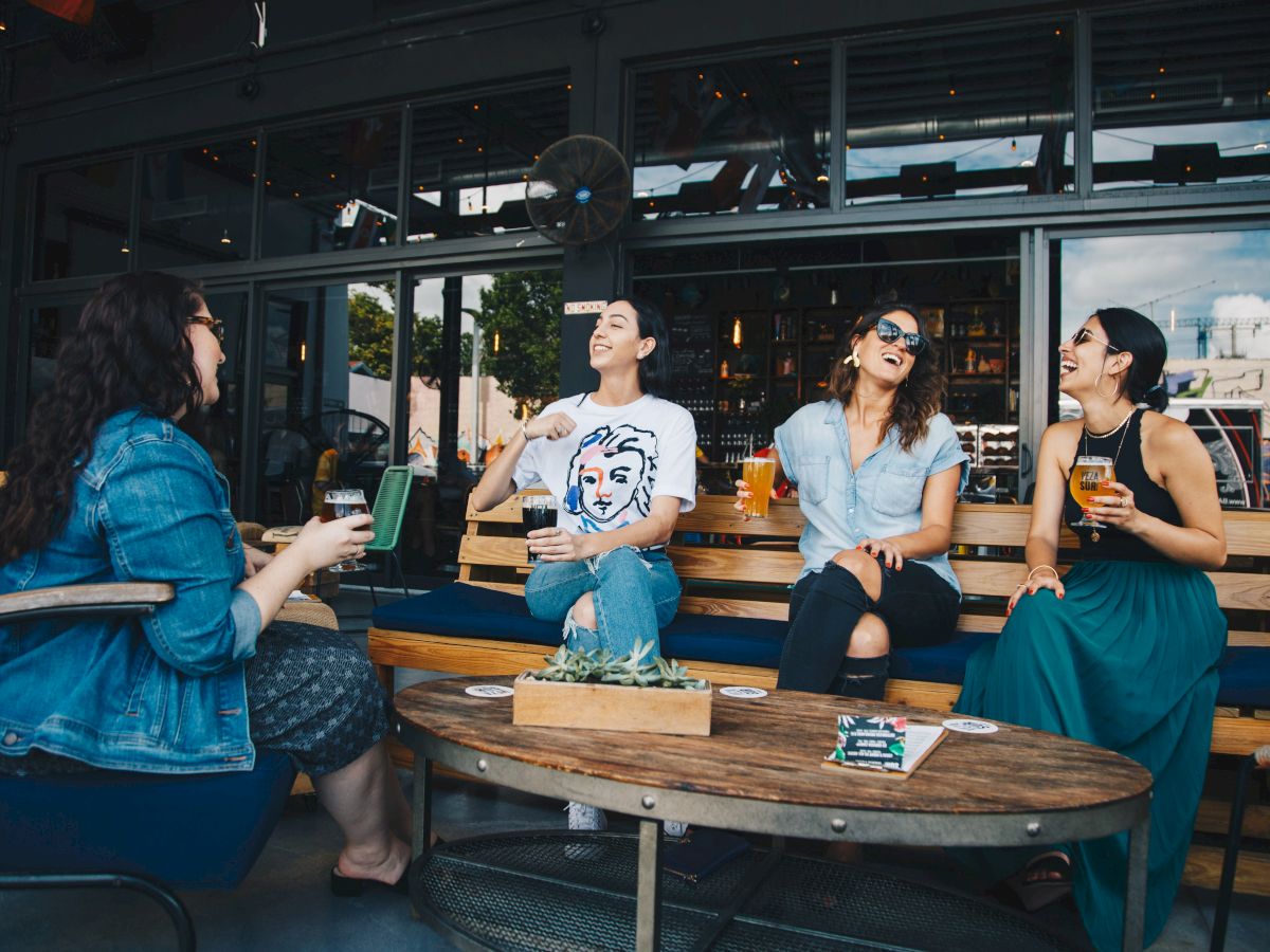 Four people sitting and laughing together at an outdoor seating area, enjoying drinks, on a sunny day, surrounding a wooden table.