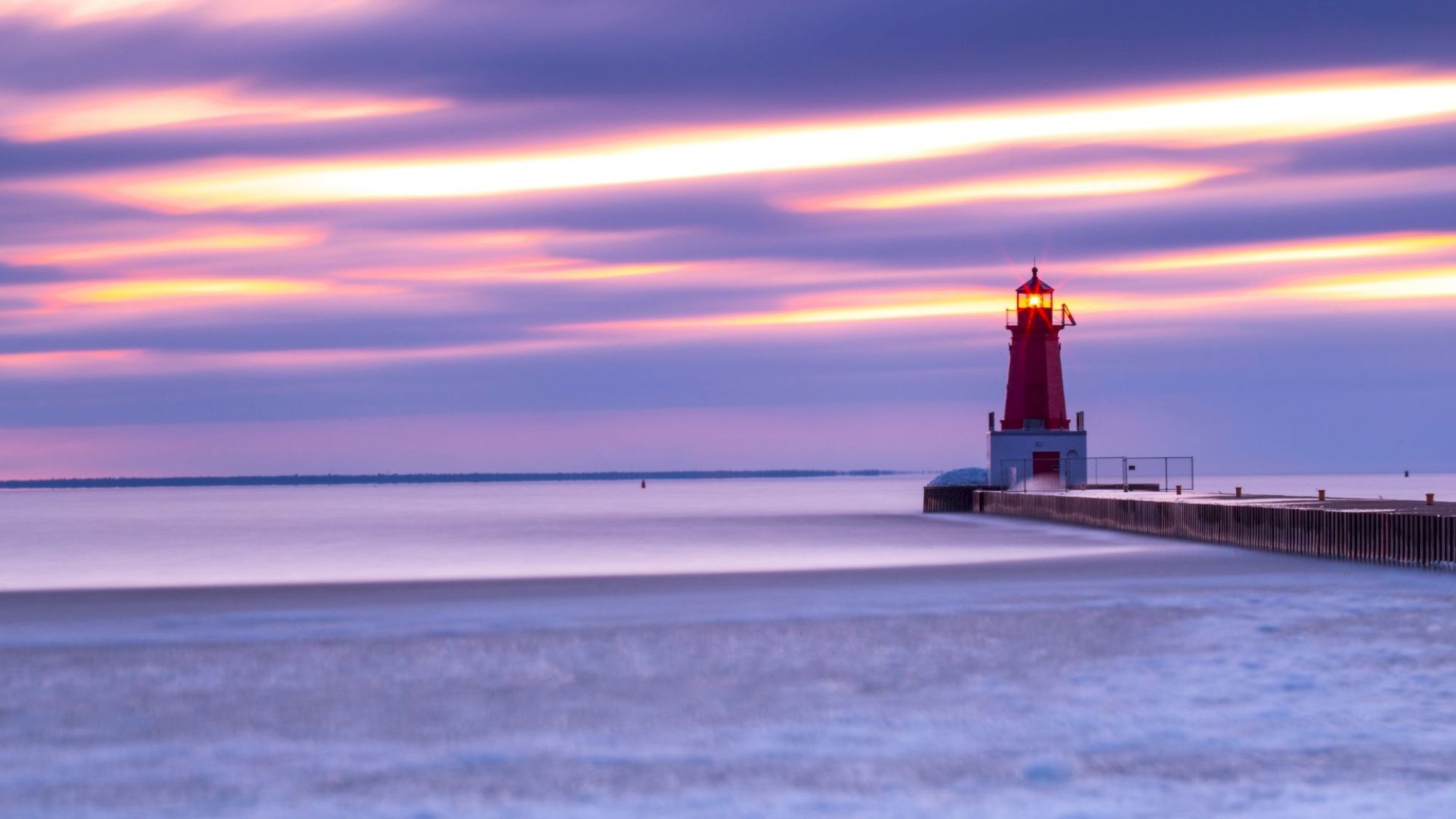 A red lighthouse stands on a pier overlooking a calm sea during sunset, with long, colorful clouds streaking across the sky.