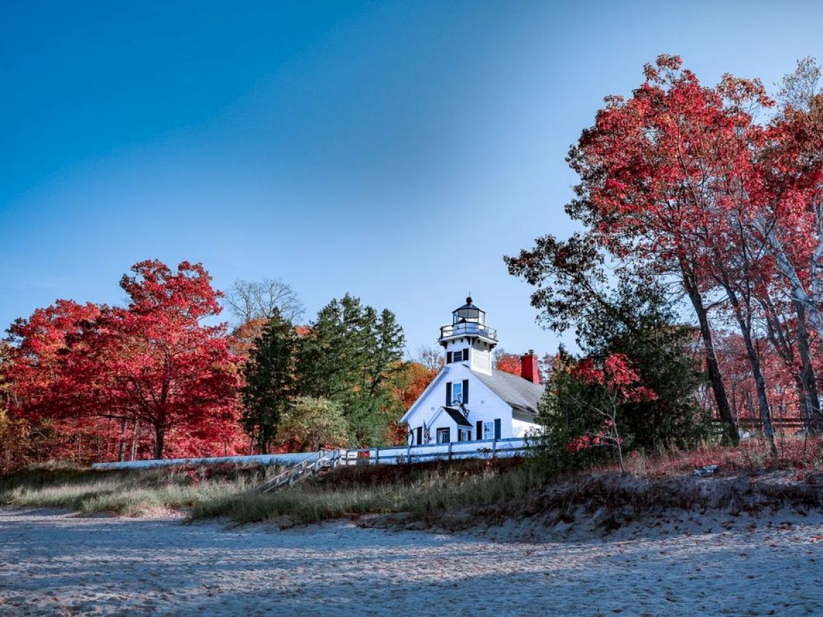 A white lighthouse is surrounded by vibrant red autumn trees under a clear blue sky, with sandy ground in the foreground, creating a serene scene.