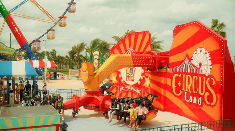 An amusement park ride with "Circus Land" signage, bright colors, and people seated and waiting for the ride to start, surrounded by other park attractions.