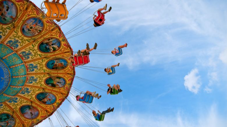 People are riding a colorful swing carousel against a blue sky. The riders are seated in chairs connected by chains, swinging outward.