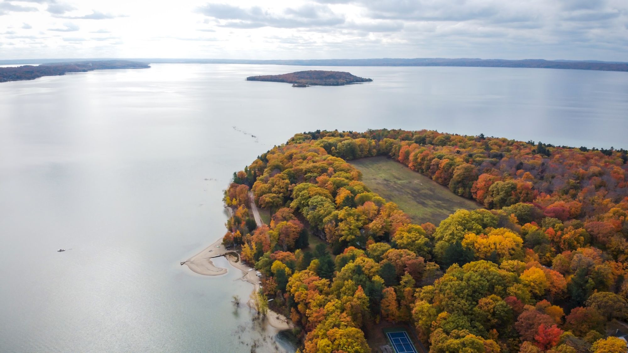 Aerial view of a peninsula covered with colorful autumn trees, surrounded by a large lake with a sandy beach and a small distant island.