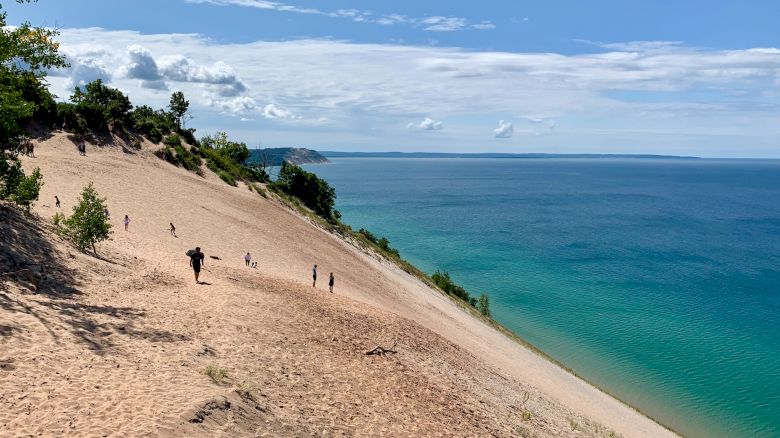 People are walking on a large sandy dune next to a blue body of water, with the sky partly cloudy and trees on the higher ground.