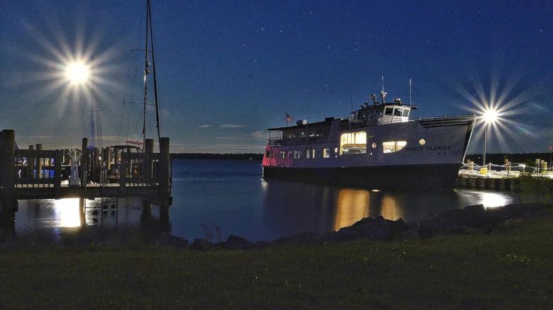 A well-lit boat docks at night with reflections shimmering on the water and starry skies above, creating a tranquil and scenic view.