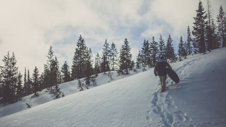 A person is trekking up a snowy hill carrying a snowboard, leaving a trail of footprints, with pine trees in the background and a cloudy sky.