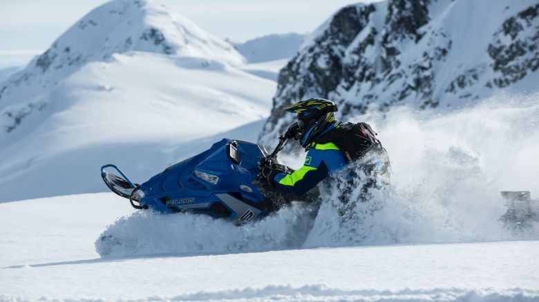 A person is riding a blue snowmobile through snowy mountains, creating a spray of snow behind them.