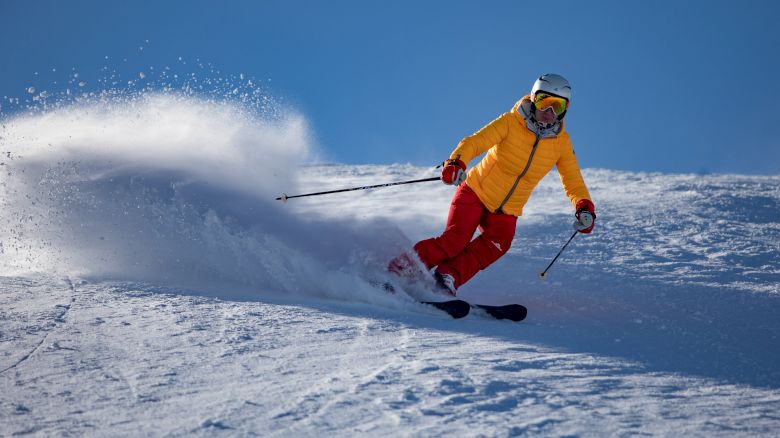 A person is skiing on snow wearing a yellow jacket and red pants, creating a spray of snow as they make a turn.