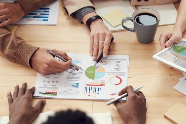 Several people analyzing charts and graphs at a meeting; there is a tablet, papers, pens, and a cup of coffee on the table.