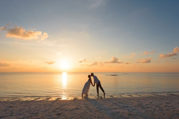 A couple is kissing on a beach at sunset with the sun setting on the horizon, surrounded by calm water and a few clouds in the sky.