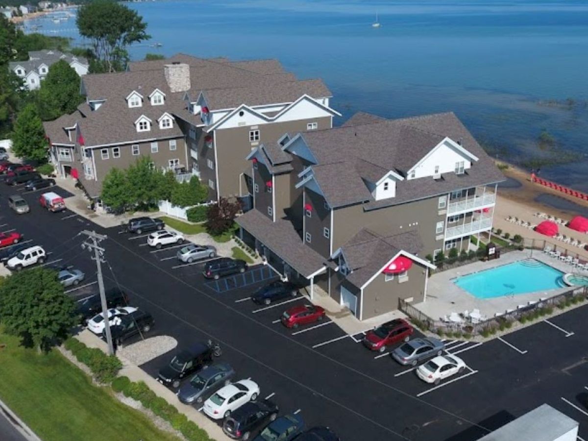 The image shows a hotel complex with a swimming pool, parking lot, and beach access beside a lake or ocean with umbrellas and lounge chairs.