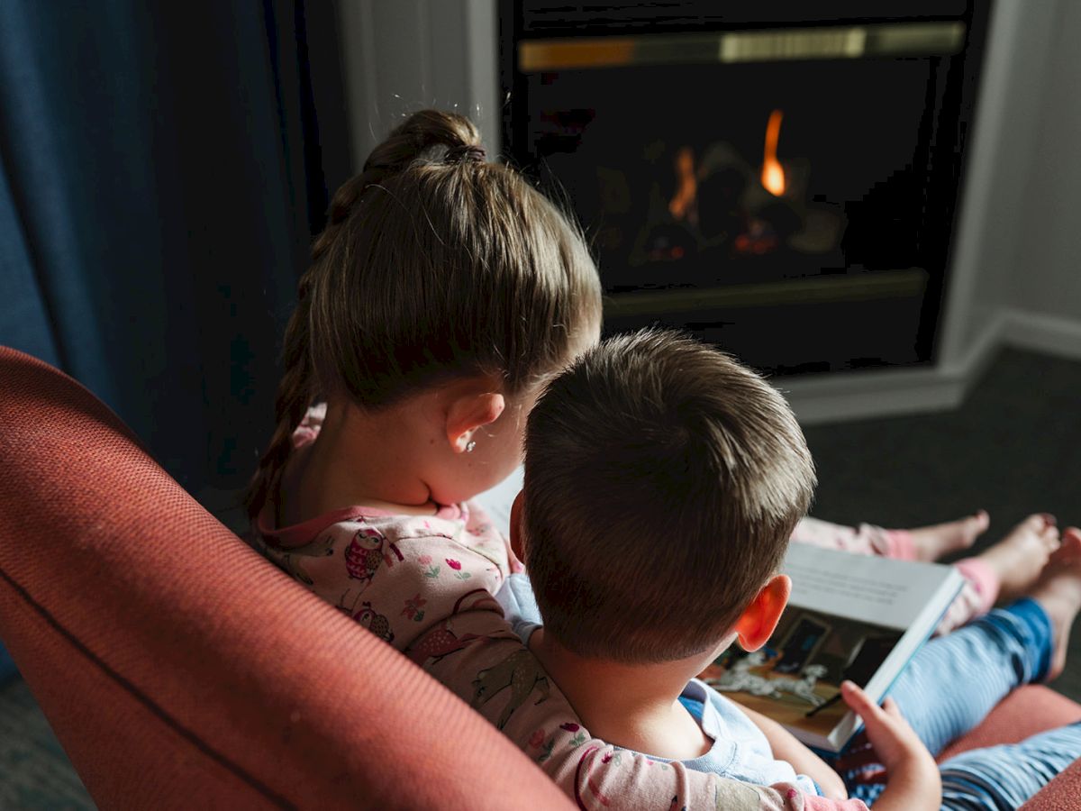 Two children sitting on a chair reading a book together, facing a lit fireplace in a cozy room.