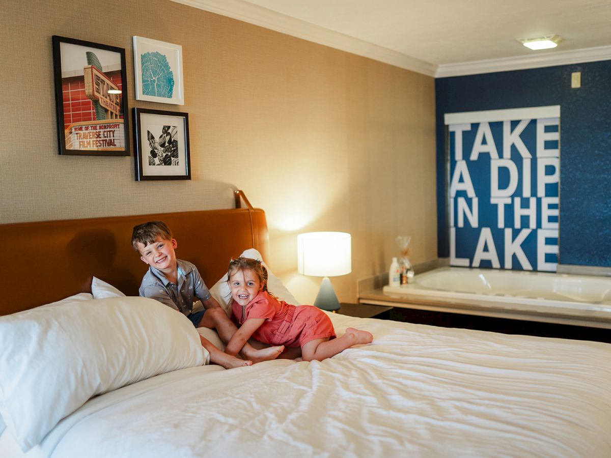 Two children play on a bed in a cozy room with art on the wall, a lamp beside the bed, and a sign in the background that reads 