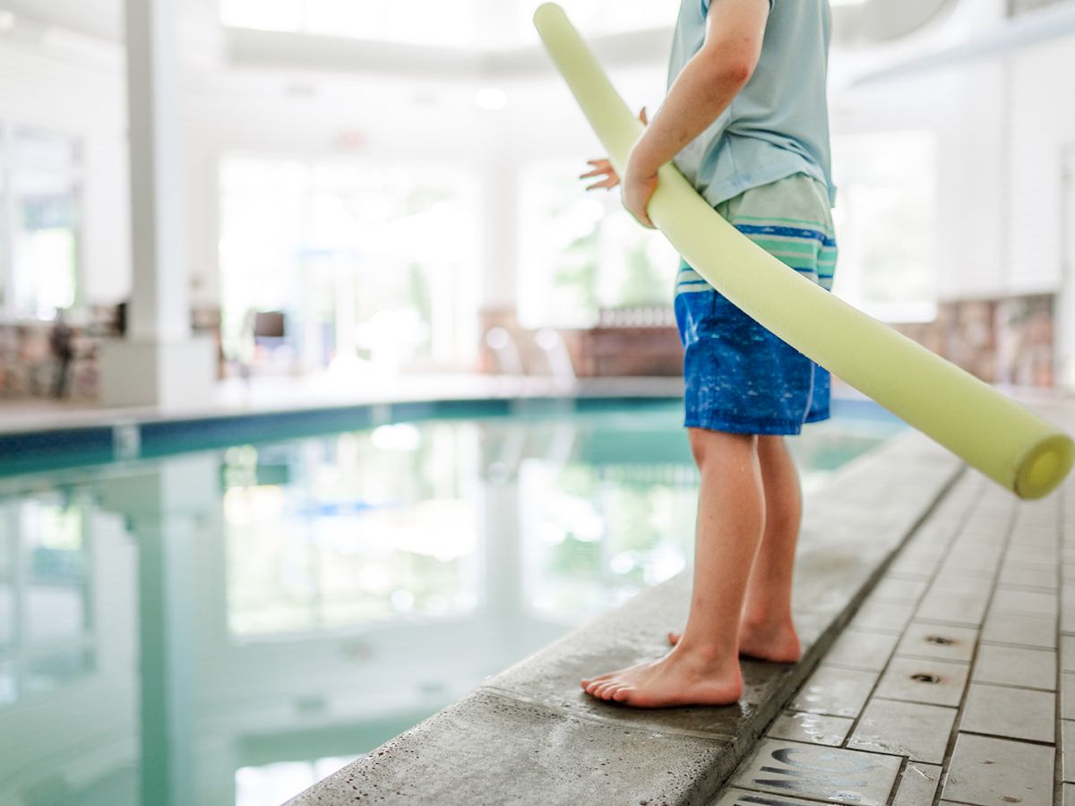 A child stands by an indoor swimming pool holding a yellow pool noodle, ready to swim.