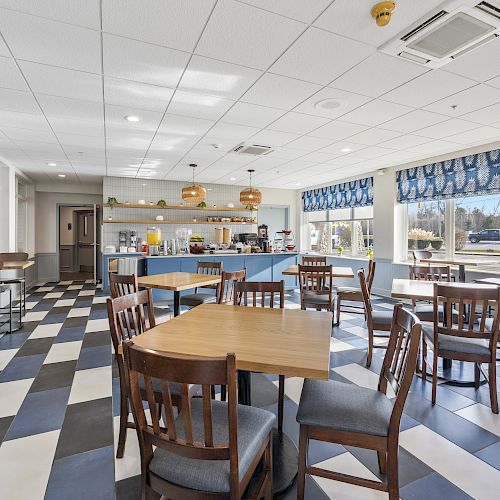 A bright, modern dining area with wooden tables and chairs, checkered flooring, and a self-service counter at the back, well-lit by large windows.