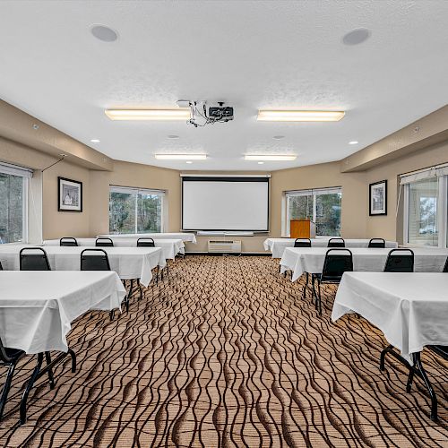 A conference room with multiple tables and chairs, white tablecloths, a projector, a screen at the front, and large windows providing natural light.