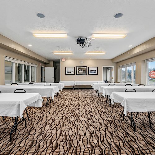 A conference room with rows of tables and chairs, white tablecloths, a projector, ceiling lights, and large windows showing the outside view.