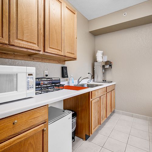 This image shows a kitchen area with wooden cabinets, microwave, sink, coffee maker, and mini fridge, on a light-colored tiled floor.