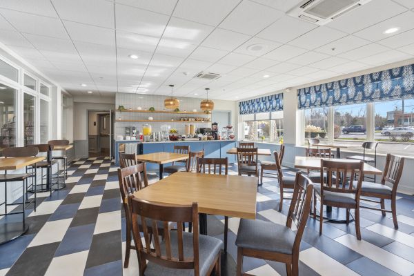 A bright, modern dining area with wooden tables and chairs, a checkered floor, large windows, and a counter with hanging lights in the background.