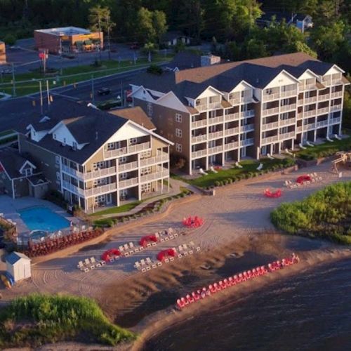 An aerial view of a waterfront resort with sandy beaches, a pool, and surrounding greenery, adjacent to a road with buildings.