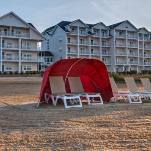 The image shows a beach area with white lounge chairs, red cabanas, and multi-story buildings in the background, resembling a resort or hotel.