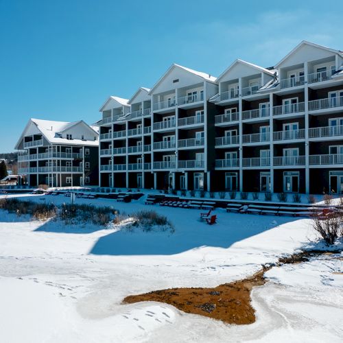 A snowy landscape with a large, multi-story building featuring balconies and a partially frozen body of water in the foreground.