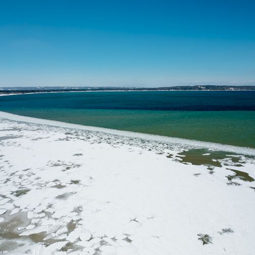 A partially frozen coastline with snow-covered land, icy patches, and a clear sky, stretching across a serene, vast water body.