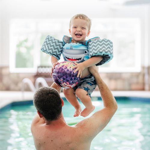 A child with floaties and a beach ball is joyfully lifted by an adult in an indoor pool.