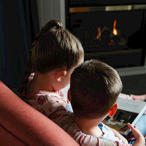 Two children are sitting on a chair, sharing a book in front of a fireplace, creating a cozy atmosphere.