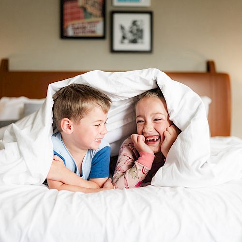 Two children are under a blanket, smiling and laughing, while lying on a bed with a headboard and framed pictures on the wall in the background.