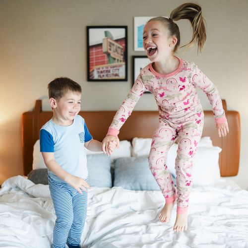 Two children are joyfully jumping on a bed in a room, wearing pajamas. The background has framed pictures and lamps on side tables.