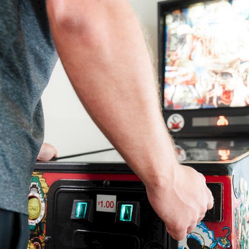 A person playing a pinball machine, focusing on the controls and colorful graphics on the machine's cabinet.