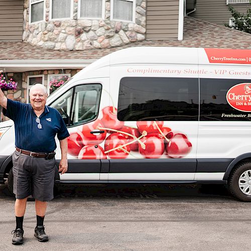 A person waves in front of a CherryTree Inn & Suites shuttle van parked by a building. The van has cherry graphics and a hotel name.