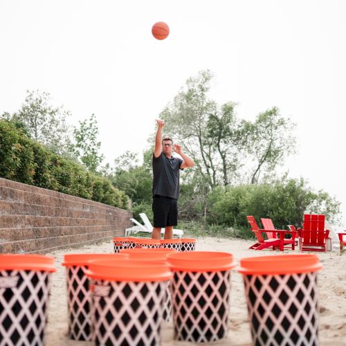 A person is playing a beach basketball game with large, orange-lidded buckets on the sand, while red chairs are placed in the background.