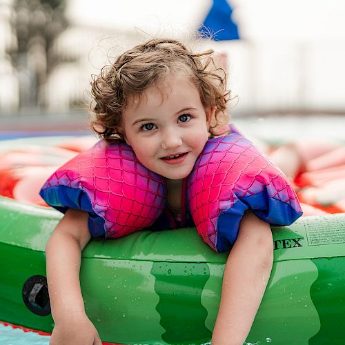 A child with curly hair is wearing bright pink and purple floaties, floating on a green inflatable in a pool, looking content.